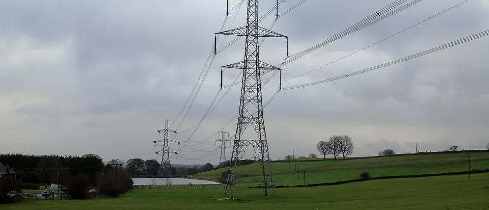 A line of high-voltage electricity pylons crossing green fields under a grey sky in England.